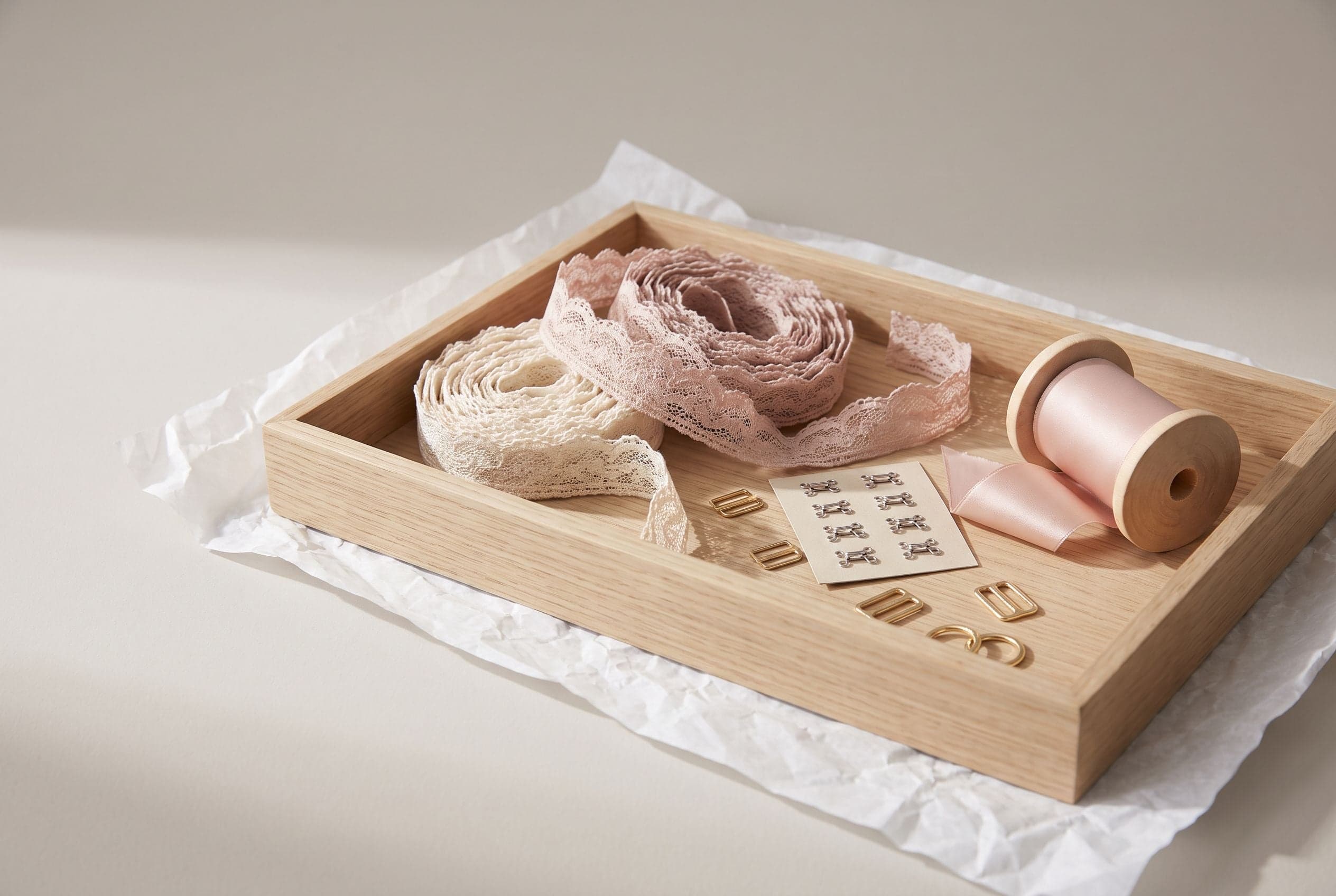 Rotoli di pizzo écru e cipria, nastro di raso e piccoli regolatori per spalline in un vassoio basso di rovere