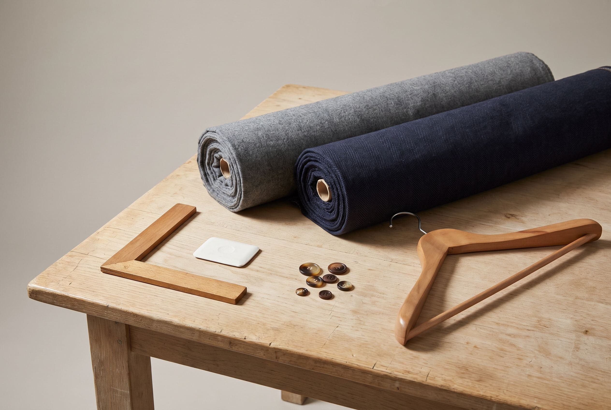 Rolls of grey flannel and navy herringbone suiting with a tailor's square, chalk, horn buttons and a wooden hanger