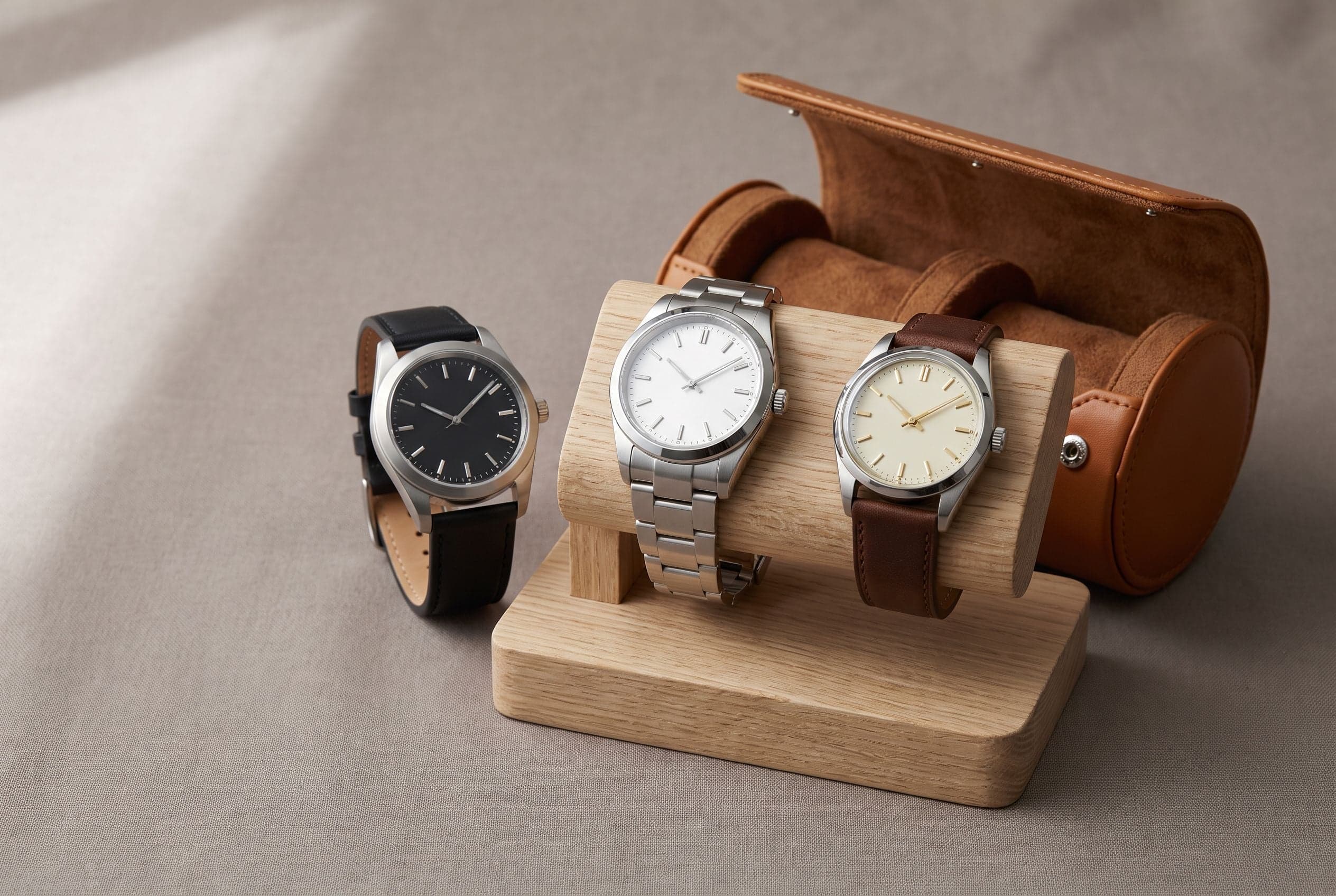 Three wristwatches with plain dials on an oak watch stand beside an open leather watch roll